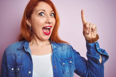 İzole edilmiş pembe arka plan üzerinde duran güzel, kızıl saçlı kadın başarılı bir fikir işaret ediyor. Heyecanlı ve mutlu. Bir numara..