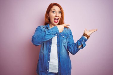 Izole pembe arka plan üzerinde duran denim gömlek giyen güzel kızıl saçlı kadın şaşırttı ve el ile sunarken kameraya gülümseyerek ve parmak ile işaret.