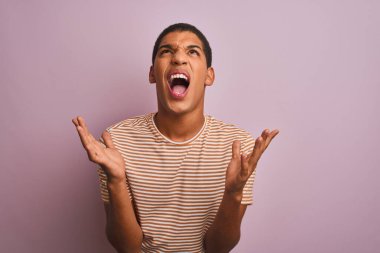 İzole edilmiş pembe arka plan üzerinde duran çizgili tişört giyen genç, yakışıklı Arap adam sinirli bir şekilde bağırıyor ve agresif bir ifadeyle kollarını kaldırıyor. Hayal kırıklığı konsepti.