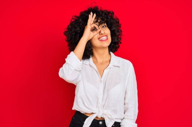 Kıvırcık saçlı, beyaz tişörtlü, izole kırmızı arka planda genç bir Arap kadın el hareketleriyle gülümsüyor ve mutlu yüzüyle parmaklarının arasından bakıyor..