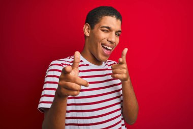 İzole edilmiş kırmızı arka plan üzerinde çizgili tişört giyen yakışıklı Arap adam mutlu ve komik yüzlü parmaklarını kameraya doğrultuyor. İyi enerji ve titreşimler.