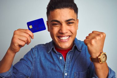 Genç Brezilyalı müşteri kredi kartı tutuyor. İzole edilmiş beyaz arka planda duruyor. Gururla bağırıyor, zaferi ve başarıyı kutluyor.