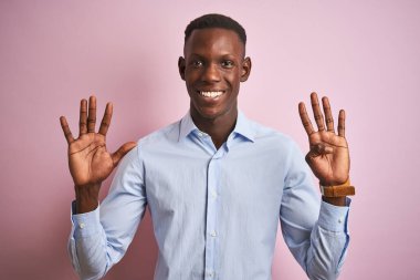 Mavi elshirt giyen Afro-Amerikalı bir adam pembe arka planda dikiliyor ve dokuz numaralı parmaklarıyla işaret ederken kendinden emin ve mutlu gülümsüyor..