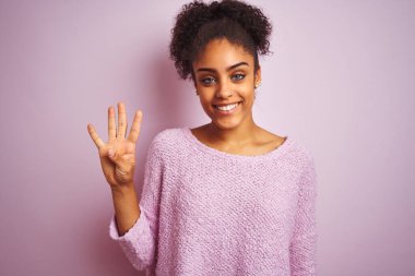 Kış süveteri giyen Afrikalı genç bir kadın pembe arka planda dikiliyor ve parmaklarını dört numarayı göstererek gülümsüyor..