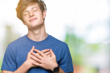 Yakışıklı delikanlı ile gülümseyen izole arka plan üzerinde mavi t-shirt giyen kapalı gözler ve yüzünde minnettar jest ile göğüs üzerinde eller. Sağlık kavramı.