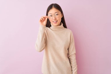 Balıkçı yaka kazak ve gözlük takan genç Çinli kadın pembe arka planda mutlu bir yüzle ayakta duruyor ve gülümsüyor dişlerini gösteren bir gülümsemeyle.