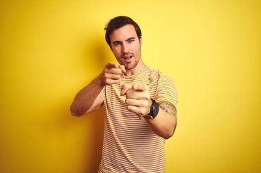 İzole edilmiş sarı arka plan üzerine çizgili tişört giyen yakışıklı genç adam mutlu ve komik yüzlü parmaklarını kameraya doğrultuyor. İyi enerji ve titreşimler.
