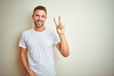 Zafer işareti yapıyor parmak gösterilen kamera seyir gülümseyen izole arka plan üzerinde rahat beyaz t-shirt giyen genç yakışıklı adam. İki numaralı.