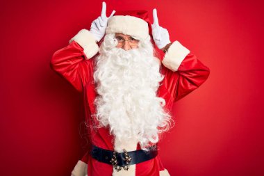 Noel Baba kostümü giymiş orta yaşlı yakışıklı bir adam izole edilmiş kırmızı arka planda duruyor komik ve çılgın pozlar veriyor parmakları kafasında tavşan kulağı, neşeli bir gülümseme