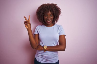 Young african afro woman wearing t-shirt standing over isolated pink background smiling with happy face winking at the camera doing victory sign. Number two.