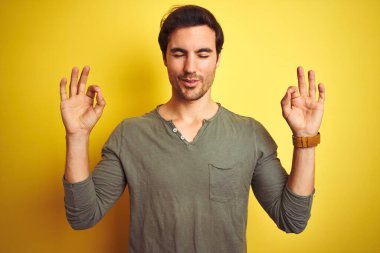 İzole edilmiş sarı arka plan üzerinde duran sıradan tişört giyen genç yakışıklı adam rahatla ve parmaklarıyla meditasyon hareketi yaparken gözleri kapalı gülümse. Yoga konsepti.