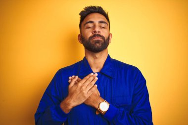 Sarı arka planda üniforma giyen yakışıklı Hintli işçi elleri göğsünde, gözleri kapalı ve yüzünde minnet dolu bir jest ile gülümsüyor. Sağlık kavramı.