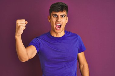 İzole edilmiş mor arka plan üzerinde duran genç Hintli adam öfkeyle bağırırken kızgın ve öfkeli bir şekilde yumruğunu kaldırıyor. Öfke ve agresif kavram.
