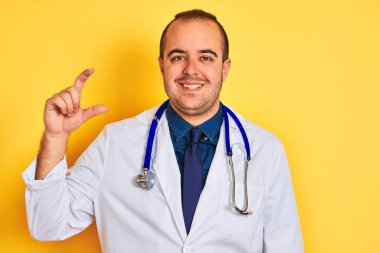 Ceket ve steteskop giyen genç doktor, izole edilmiş sarı arka planda duruyor gülümsüyor ve el hareketleriyle küçük boy işareti yapıyor, parmakları bakıyor ve kameraya bakıyor. Ölçü kavramı.