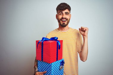 Dövmeli genç adam, elinde doğum günü hediyeleri ile izole edilmiş beyaz arka planda durup gururla haykırıyor zaferi ve başarıyı kutluyor çok heyecanlı, coşkulu bir duygu.