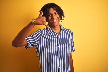Rastgele çizgili tişört giyen Afro adam izole edilmiş sarı arka planda gülümseyen el ve parmaklarıyla telefon hareketi yapan telefonda konuşan adam gibi. İletişim kavramları.