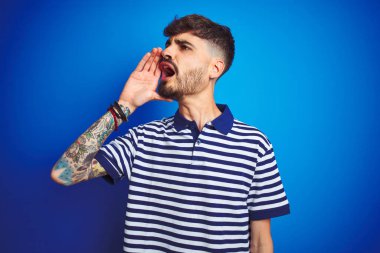 İzole edilmiş mavi arka planda duran çizgili polo giyen genç adam el ele tutuşarak bağırıyor ve bağırıyor. İletişim konsepti.