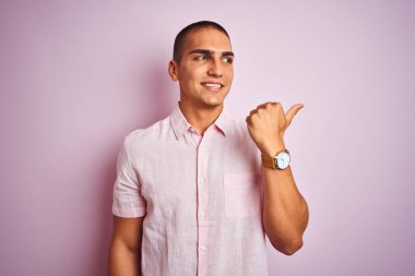 Pembe izole edilmiş arka planda zarif gömlekler giyen yakışıklı bir genç. Gülümsüyor, gülümsüyor ve başparmağıyla yan tarafa bakıyor..