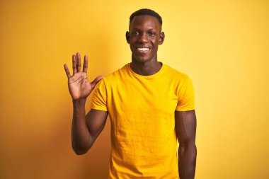 Genç Afro-Amerikalı bir adam gündelik tişört giyip izole edilmiş sarı arka planda dikiliyor ve beş numarayı işaret ederek kendinden emin ve mutlu gülümsüyor..