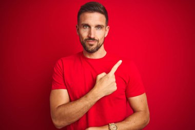 Kırmızı izole edilmiş arka plan üzerine gündelik tişört giymiş genç yakışıklı bir adam. El parmağı yanda, ciddi ve sakin bir yüz ifadesi sergiliyor.