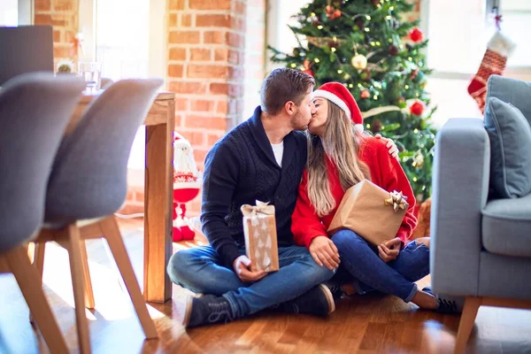 年轻漂亮的夫妇面带微笑 既快乐又自信 坐在地板上拿着礼物 在家里的圣诞树旁拥抱和亲吻 — 图库照片