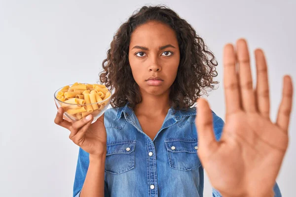 若いですブラジル人女性保持ボウルとともにMacaroniパスタ上の隔離された白い背景でオープン手を行います停止標識で真剣で自信を持って式 防衛ジェスチャー — ストック写真