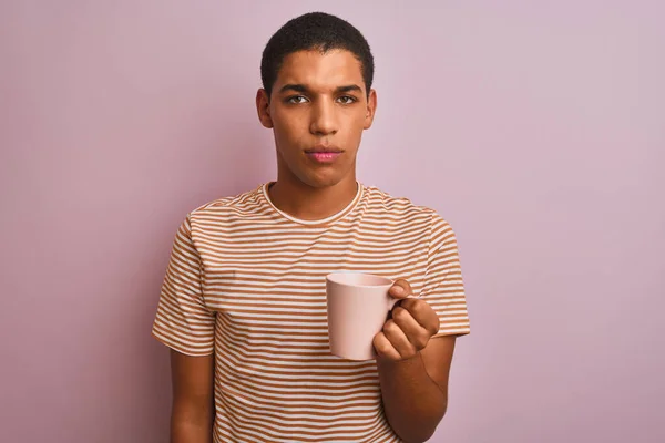 英俊的阿拉伯男子 身穿条纹T恤衫 带着淡淡的粉色背景喝咖啡 脸上带着自信的表情 一副严肃严肃的表情 — 图库照片