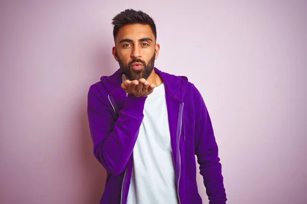 Young indian man wearing purple sweatshirt standing over isolated pink background looking at the camera blowing a kiss with hand on air being lovely and sexy. Love expression.