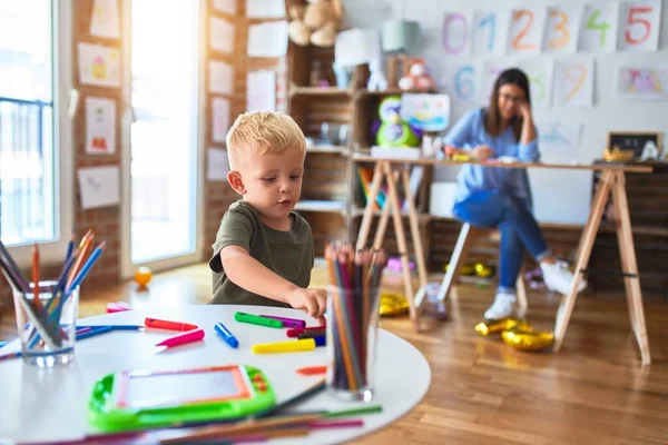 若い白人の子供は教師と一緒に学校で遊んでいる 母と息子は遊び場で色鉛筆で絵を描く若い女性は机の上に座っている — ストック写真