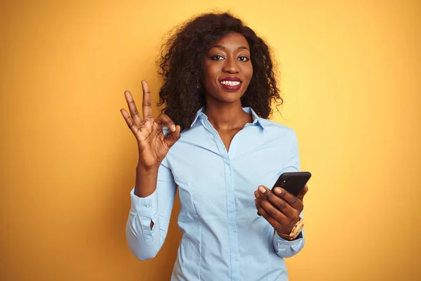 年轻的非洲裔美国女人用智能手机站在孤立的黄色背景上 手握手签 很好的符号 — 图库照片
