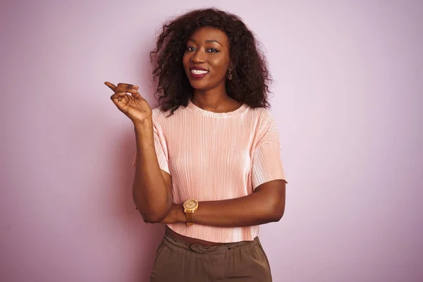 Young african american woman wearing t-shirt standing over isolated pink background with a big smile on face, pointing with hand and finger to the side looking at the camera.