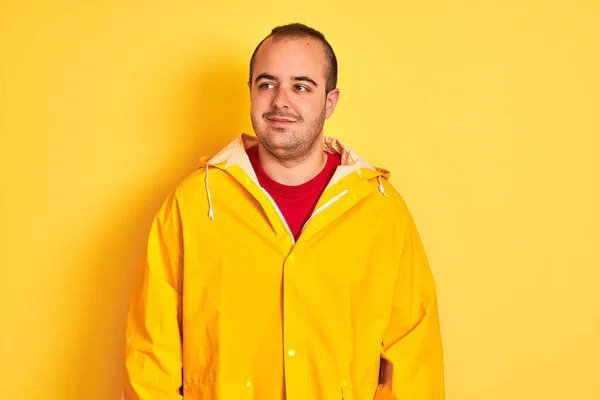 若い男がレインコートを着て孤立した黄色の背景の上に立って笑顔側を見て 離れて考えて見つめて — ストック写真