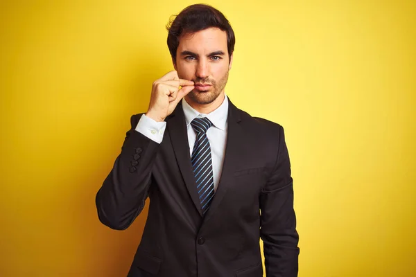 Young handsome businessman wearing suit and tie standing over isolated yellow background mouth and lips shut as zip with fingers. Secret and silent, taboo talking