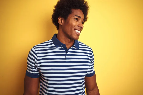 American man with afro hair wearing navy striped polo standing over isolated yellow background looking away to side with smile on face, natural expression. Laughing confident.