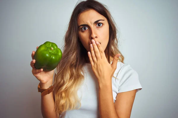 Young beautiful woman holding green pepper over white isolated background cover mouth with hand shocked with shame for mistake, expression of fear, scared in silence, secret concept