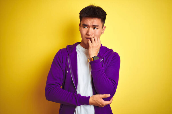 Young asian chinese man wearing purple sweatshirt standing over isolated yellow background looking stressed and nervous with hands on mouth biting nails. Anxiety problem.