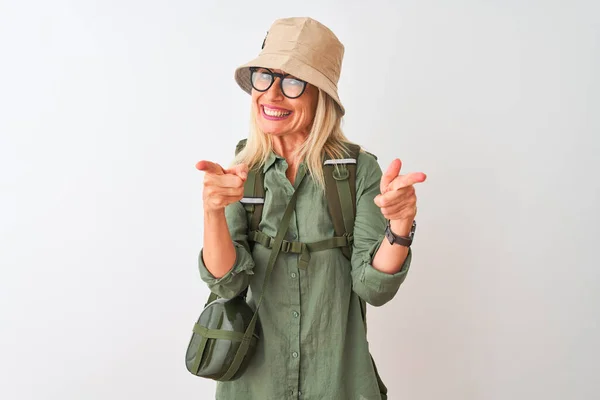 Middle age hiker woman wearing backpack hat canteen glasses over isolated white background pointing fingers to camera with happy and funny face. Good energy and vibes.