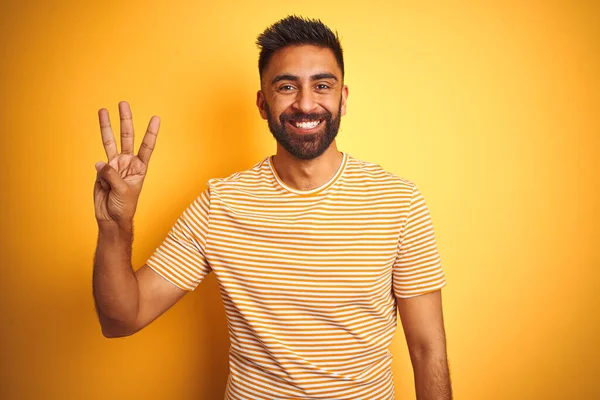 Young indian man wearing t-shirt standing over isolated yellow background showing and pointing up with fingers number three while smiling confident and happy.