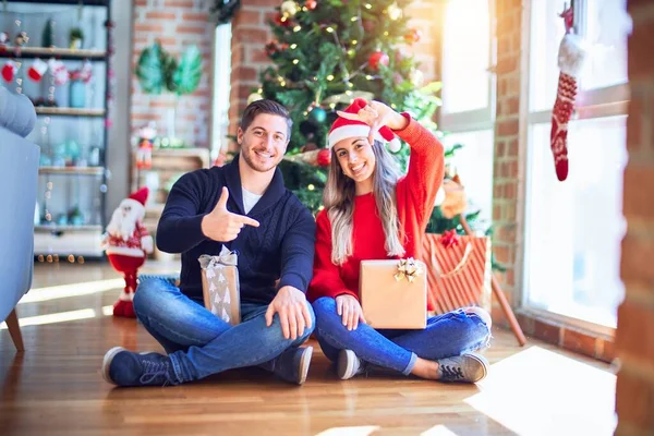 年轻夫妇戴着圣诞老人帽 坐在自家圣诞树下的地板上 笑容满面 手指手画脚 喜形于色 创意和摄影概念 — 图库照片