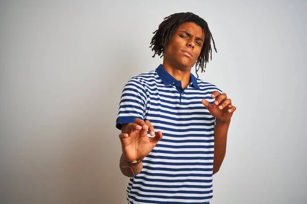 Afro man with dreadlocks wearing striped blue polo standing over isolated white background disgusted expression, displeased and fearful doing disgust face because aversion reaction. With hands raised. Annoying concept.