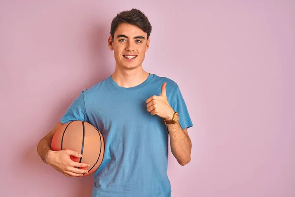 Teenager boy holding professional basket ball over isolated pink background happy with big smile doing ok sign, thumb up with fingers, excellent sign