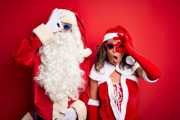 中年夫妇穿着圣诞老人服装 戴着太阳镜 站在孤立的红色背景上 摆出一副很好的姿势 脸上带着惊讶的表情 用手指看着对方 难以置信的表情 — 图库照片