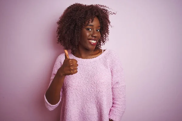 Young african afro woman wearing sweater standing over isolated pink background doing happy thumbs up gesture with hand. Approving expression looking at the camera showing success.