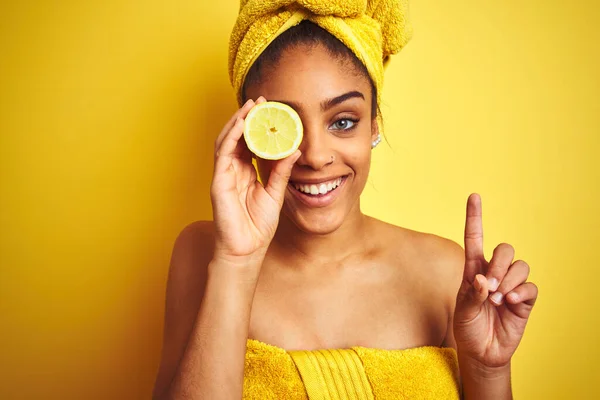 シャワー後のタオルを身に着けているアフロ女性は幸せな顔 ナンバーワンでアイデアや質問を指差す指で驚いて隔離された黄色の背景にスライスレモンを保持 — ストック写真