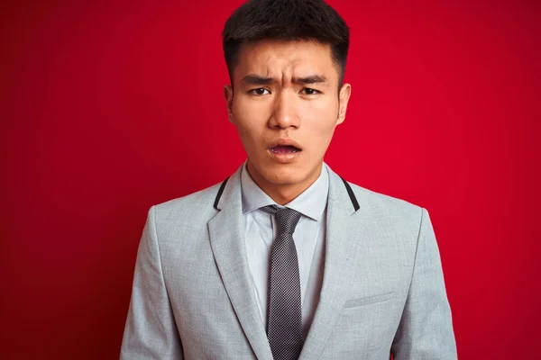 Asian chinese businessman wearing grey jacket and tie standing over isolated red background afraid and shocked with surprise expression, fear and excited face.