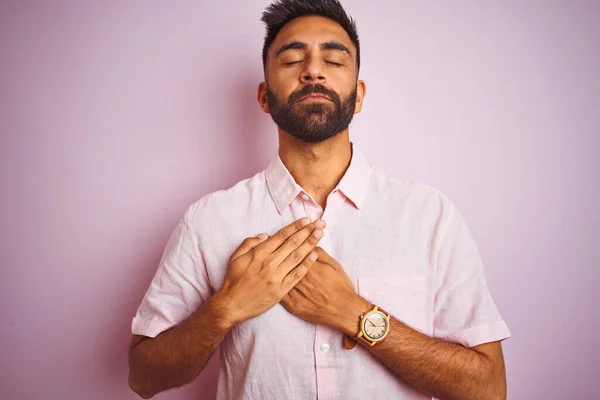 Young indian man wearing casual shirt standing over isolated pink background smiling with hands on chest with closed eyes and grateful gesture on face. Health concept.