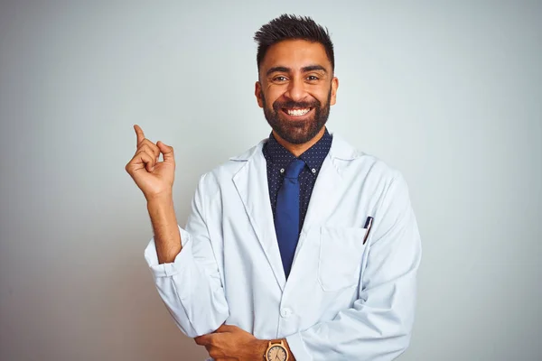 Young indian doctor man standing over isolated white background with a big smile on face, pointing with hand and finger to the side looking at the camera.