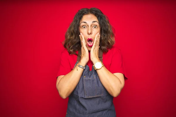 Middle age senior woman wearing apron uniform over red isolated background afraid and shocked, surprise and amazed expression with hands on face