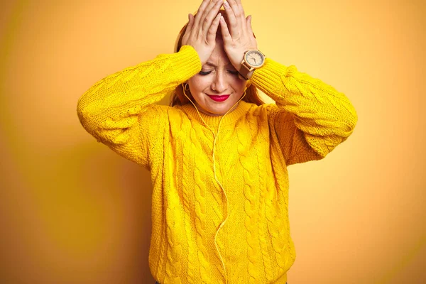 Young redhead woman listening to music using headphones over yellow isolated background suffering from headache desperate and stressed because pain and migraine. Hands on head.