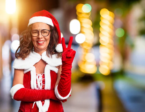 中年妇女穿着圣诞老人的服装 喜形于色地微笑着 在镜头前眨眼示意胜利 第二点 — 图库照片
